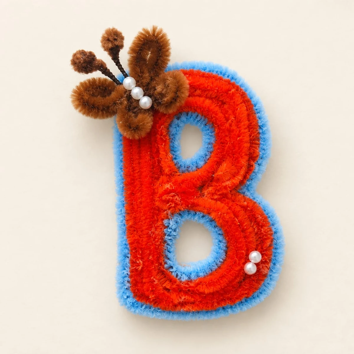 Butterfly Initial B: A bold orange B outlined in soft blue, with a tiny butterfly and pearl-like accents.